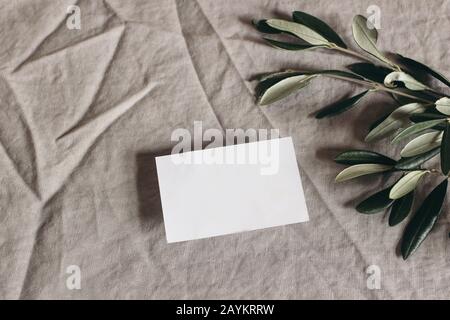 Moody feminine Hochzeitstatikerei Mock-up-Szene. Leerer Gruß, Visitenkarte und grüne olivgrüne Blätter, Äste auf Leinen-Tischdecke-Hintergrund, flach l. Stockfoto