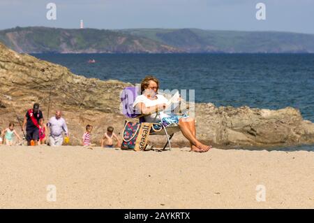 Alleinstehende Frau, die auf einem Strand in einem Liegestuhl sitzt und eine Zeitung liest Stockfoto