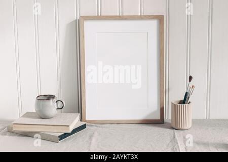 Leeres Holzrahmen-Mockup mit Farbbürsten, Bleistiften in Keramikhalter, Tasse Kaffee und Büchern auf Leinentisch. Künstlerische Szene. Kreativtisch B. Stockfoto