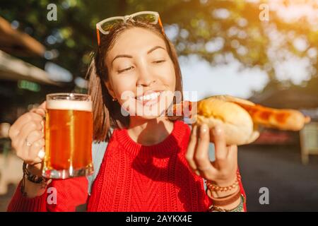 Junge fröhliche asien-reisende Frau trinkt Bierbecher mit Hotdog in Deutschland, Bier- und Food-Festival-Konzept Stockfoto