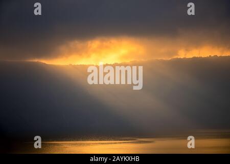 Schwere Wolken über dem felsigen Ufer. Die Sonnenstrahlen brechen kaum durch, beleuchten kleine Häuser und die Wasseroberfläche Stockfoto