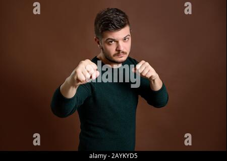 Der junge Mann mit braunen Haaren zeigt beiläufig seine Fäuste und wird wütend auf den isolierten braunen Hintergrund Stockfoto