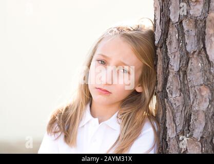 Ein wunderschönes, junges, blondes, blauäugiges Mädchen mit traurigen Augen und einem melancholischen Blick auf ihr Gesicht, das ein weißes Hemd trägt und sich an den Stamm von lehnt Stockfoto