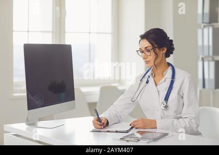 Selbstbewusste Frau Ärztin Kinderärztin schreibt in einer Zwischenablage, die an einem Tisch in einem weißen Büro des Krankenhauses sitzt Stockfoto