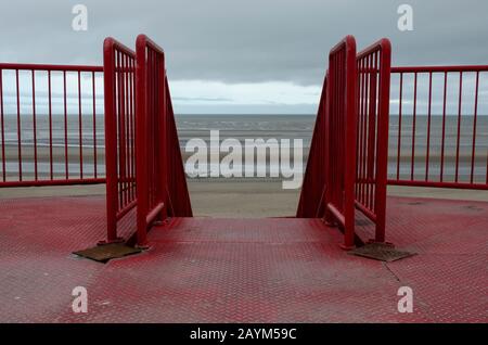 Verwittertes, rostiges und getragenes rotes Metall-Geländer mit Stufen, die an einem grauen Wintertag in Richtung Strand führen Stockfoto