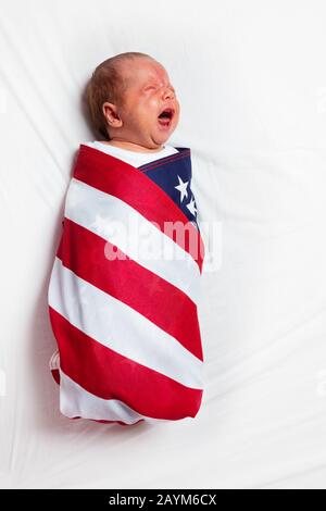 Porträt des kleinen weinenden Säuglingsjungen, der in die amerikanische Flagge gehüllt wurde, die auf dem Bettlaken aufliegt Stockfoto