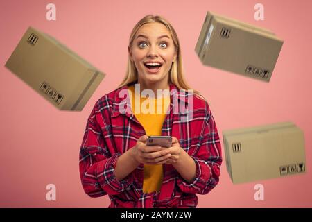 Blondes süßes Mädchen erhält Priorität schnelle Kästen aus Online-Shop-Auftrag. Überraschter und verblüffter Ausdruck. Rosafarbener Hintergrund Stockfoto