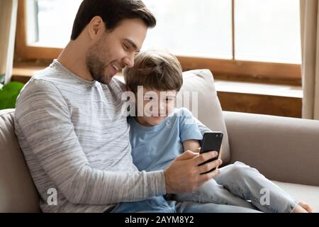 Fröhlicher junger Vater, der kleinen Sohn kuschelt und lustige mobile Anwendungen zeigt. Stockfoto