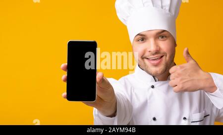 Küchenchef Zeigt Smartphone-Bildschirm Gestikulierend Thumbs-Up Über Gelbem Hintergrund Stockfoto