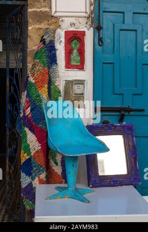 Hellblauer Stuhl, die gleiche wie die Farbe der Tür im Hintergrund, auf der Straße vor einem Geschäft ausgesetzt.Teppich und Rahmen. Essaouira, Marokko Stockfoto