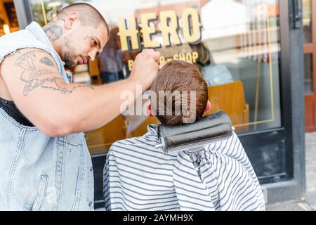 12. MAI 2018, SLOWAKEI, BRATISLAVA: Brutal aussehender barber mit vielen Tätowierungen rasiert bei heißem Sommerwetter einen Klientelmann im Freien Stockfoto