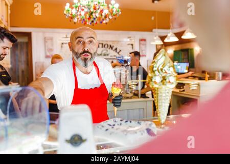 12. MAI 2018, SLOWAKEI, BRATISLAVA: Charismatischer und gutaussehender Koch mit Bart verkauft Eis an Käufer im Gelato-Laden Stockfoto