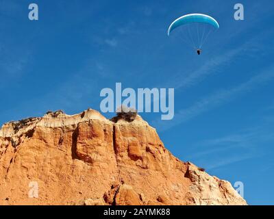 Ein Mann fliegt in seinem Fallschirm über rote Klippen in Albufeira an der Küste der Algarve in Portugal Stockfoto