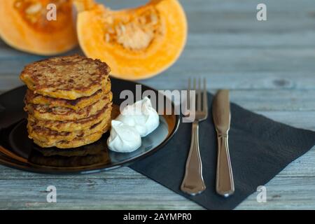 Pfannkuchen mit natürlichem Yougurt auf schwarzem Teller, Gabel, Messer, PapierServiette serviert auf grauem Holztisch, geschnittene Stücke und orangefarbener Kürbis auf dem Hintergrund Stockfoto