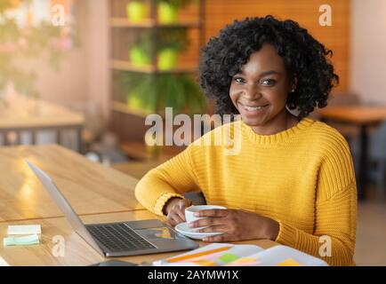 Portrait der schwarzen jungen fröhlichen Dame mit Laptop in der Cafeteria Stockfoto