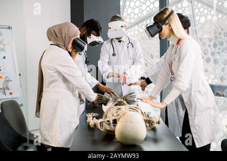 Erfahrener, erfahrener, männlicher Arzt verwendet VR-Brille und menschliches Skelett für den Unterricht von Studenten. Gruppen von multiethnischen Studenten oder Praktikanten studieren Menschen Stockfoto