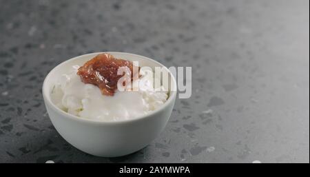 Fügen Sie Feigenmarmalade zu Hüttenkäse in weißer Schüssel auf Betonoberfläche hinzu, breites Foto Stockfoto