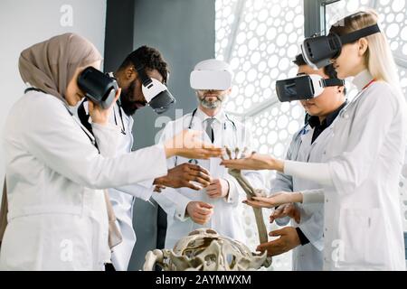 Gruppe multiethnischer Medizinstudenten oder Ärzte und reifer männlicher Professor, der die Anatomie des menschlichen Skeletts im Klassenzimmer mit einem VR-Brillen-Headset untersucht Stockfoto