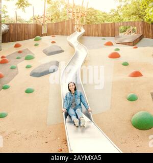 Eine infantile und glückliche Frau, die auf einer Kinderrutsche auf einem Spielplatz sitzt Stockfoto