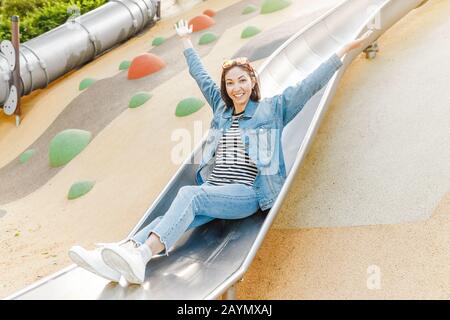 Eine infantile und glückliche Frau, die auf einer Kinderrutsche auf einem Spielplatz sitzt Stockfoto