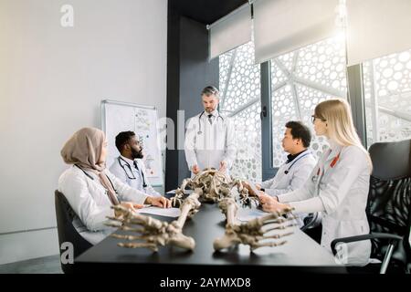Junge multiethnische Medizinstudenten zum Anatomieunterricht im modernen Klassenzimmer. Professor für männlichen Mittelalter-Arzt, der Anatomie mit menschlichem Skelett lehrt Stockfoto