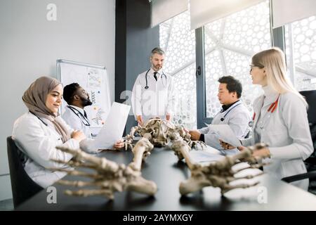 Gutaussehender Arzt im mittleren Alter, Anatomielehrer, der die Anatomie der Knochen anhand des Skelettmodells für verschiedene Schüler in weißen Mänteln erklärt. Medizin, Bildung Stockfoto