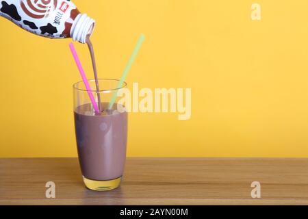 Gießen einer Muller Frijj Schokoladenmilch in ein halbes Pint Spottglas mit zwei Strohhalmen auf Holztisch mit Copyspace auf gelbem Hintergrund Stockfoto