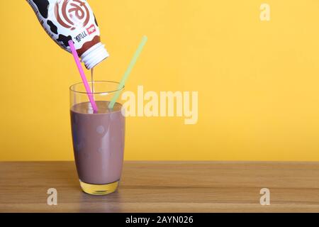 Gießen einer Muller Frijj Schokoladenmilch in ein halbes Pint Spottglas mit zwei Strohhalmen auf Holztisch mit Copyspace auf gelbem Hintergrund Stockfoto