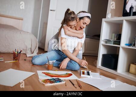 Lustige kleine Mädchen umarmt ihre Mutter, während sie ein Bild auf dem Boden malt, aufgeregte Frau mit weit offenem Mund hilft ihrer Tochter Stockfoto