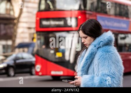 Ein einsam attraktives Weibchen in modischem furchtblauem Mantel, das sich auf ihr Handy mit Verkehr im Hintergrund konzentriert Stockfoto