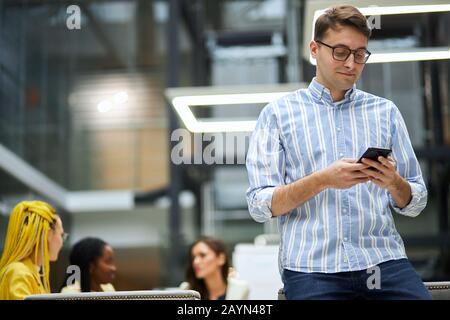 Stylieh junger Mann in Modetrikot auf den Schreibtisch gelehnt, am Rand des Tisches sitzend, Foto an Kunden, Freundin, angenehme Konversation senden Stockfoto