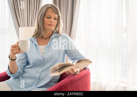 Reife Frau, die sich mit einem Buch in ihrer städtischen Wohnung erholt Stockfoto