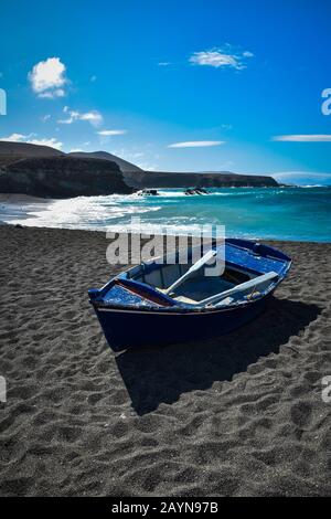 Holzboot am schwarzen Sandstrand, türkisfarbenem Ozean und schwarzen vulkanischen Klippen im Hintergrund. Blauer Himmel und kleine Wolken. Ajuy, Fuerteventura Stockfoto