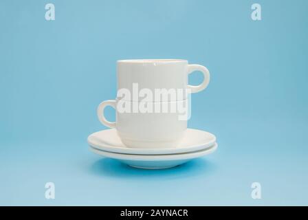 Zwei weiße Porzellanbecher und Untertasse, übereinander auf hellblauem Grund. Seitenansicht Stockfoto