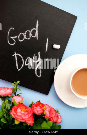 Konzeption guten Morgen. Schwarze Kreideplatte mit der Aufschrift "guten Tag", eine Tasse rosafarbenen Kaffeesärg auf hellblauem Grund. Draufsicht. Flaches Lay Stockfoto