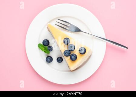 Käsekuchen mit Blaubeeren auf rosa Hintergrund. Draufsicht. Dessert des Mädchens. Klassischer einfacher New Yorker Käsekuchen auf weißem Teller Stockfoto