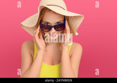 Attraktive, schicke junge Frau im Sommer-Outfit mit einem großen Floppy-Sonnenhut, der mit einem freundlichen Lächeln über die Oberseite ihrer Sonnenbrille an der Kamera schält Stockfoto