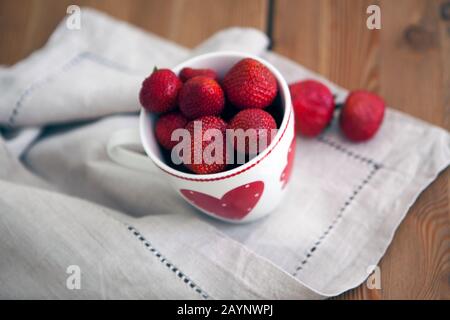 Frische reife rote Erdbeeren in einem Becher und rauem Tuch auf Holzgrund, natürliche rustikale Speisen. Rote Sommererdbeeren auf einem weißen Becher mit Herzen Stockfoto