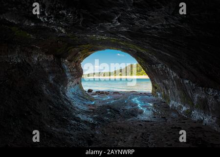 Blick von innen auf eine Meereshöhle. Blauer Himmel, Wasser, Strand und Bäume im Freien. Die Wände der Höhle sind nass und reflektieren Licht von außen. Stockfoto