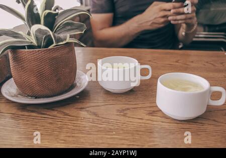 Konzeptfoto mit zwei Tassen Tee und einer Person, die das Telefon verwendet, um die Auswirkungen von Technologie, sozialen Medien und dem Internet im Alltag zu zeigen Stockfoto