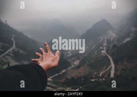 Eine Hand, die zu den nebligen Bergen und kurvenreichen Straßen von ha giang Loop in Vietnam reicht und ein filmisches und stimmungsvolles Reisefoto zeigt Stockfoto