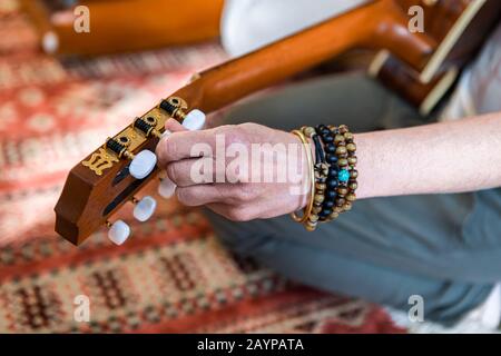 Nahaufnahme der menschlichen Hände, die klassische Gitarrensaiten so stimmen, dass sie heilige Kirtan-Musik zur Meditation spielen, während der Mann auf dem Fußbodenteppich sitzt Stockfoto