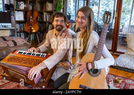 Junger schamanischer Mann und Frau, die klassische Gitarre und Harmonium spielen, während sie nach der Meditationssitzung die Kamera auf dem Bodenteppich sehen Stockfoto