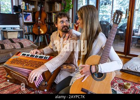 Junger schamanischer Mann und Frau, die klassische Gitarre und Harmonium spielen, während sie sich nach der Meditationssitzung auf dem Fußbodenteppich angucken Stockfoto