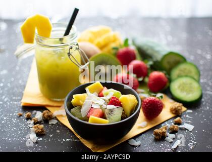 Gesundes Frühstück: Knusprige Müsli mit frischen Erdbeeren, Kiwi, Mango und Kokosflocken mit köstlichen grünen Smoothie im Mixbecher Stockfoto
