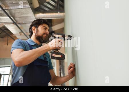Tiefwinkelporträt bärtiger Bauarbeiter bohrt Wand, während Haus allein renoviert wird, Kopierraum Stockfoto