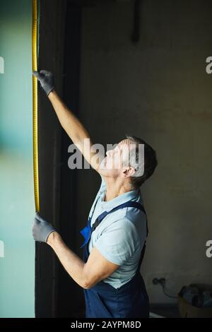 Seitenansicht von leitenden Bauarbeitern, die Wand mit Band messen, während sie das Haus renovieren, Kopierraum Stockfoto