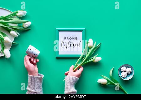 Hände mit Kaffeetasse und weißen Frühlings-Tulpen, Blumenstrauß auf Platte und Marshmallow Süßigkeiten. Flaches Layout mit grünem Hintergrund, Draufsicht, Rahmen mit Text Stockfoto
