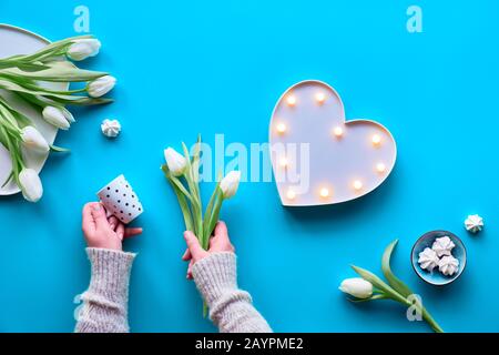 Beleuchtetes Herzlichtbrett. Kaffeetasse mit Polka-Punkten und weißen Tulpen in den Händen. Frühlings-Tulpen, mehr Blumen auf Platte und Marschlauchbonbons. Valen Stockfoto