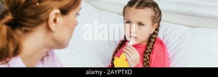 Panoramaaufnahme der traurigen kranken Tochter, die Fieber mit Thermometer misst, während sie im Bett mit Mutter sitzt Stockfoto
